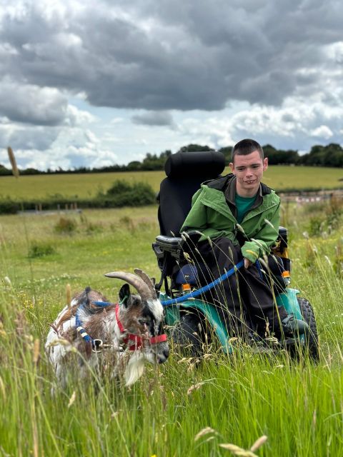 A photograph of a person we support in an electric wheelchair with a goat in the field at Blossom Barns